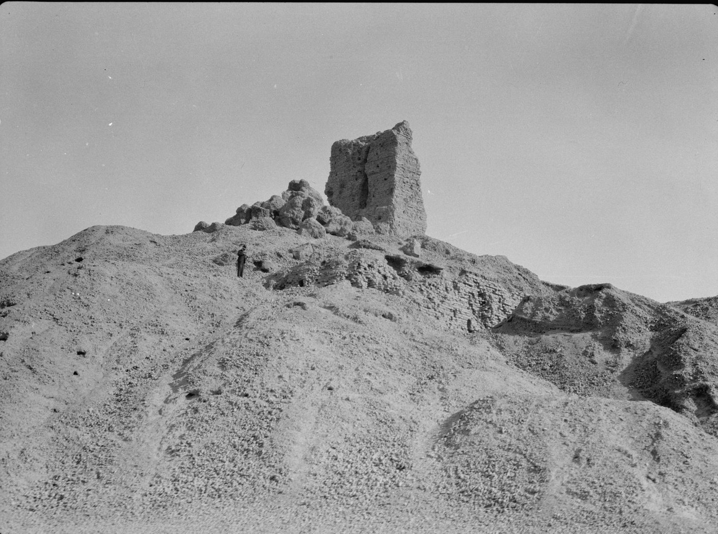 A large mound of collapsed mudbrick in the Iraqi desert with one ruined wall still standing on top, a man on the slope for scale
