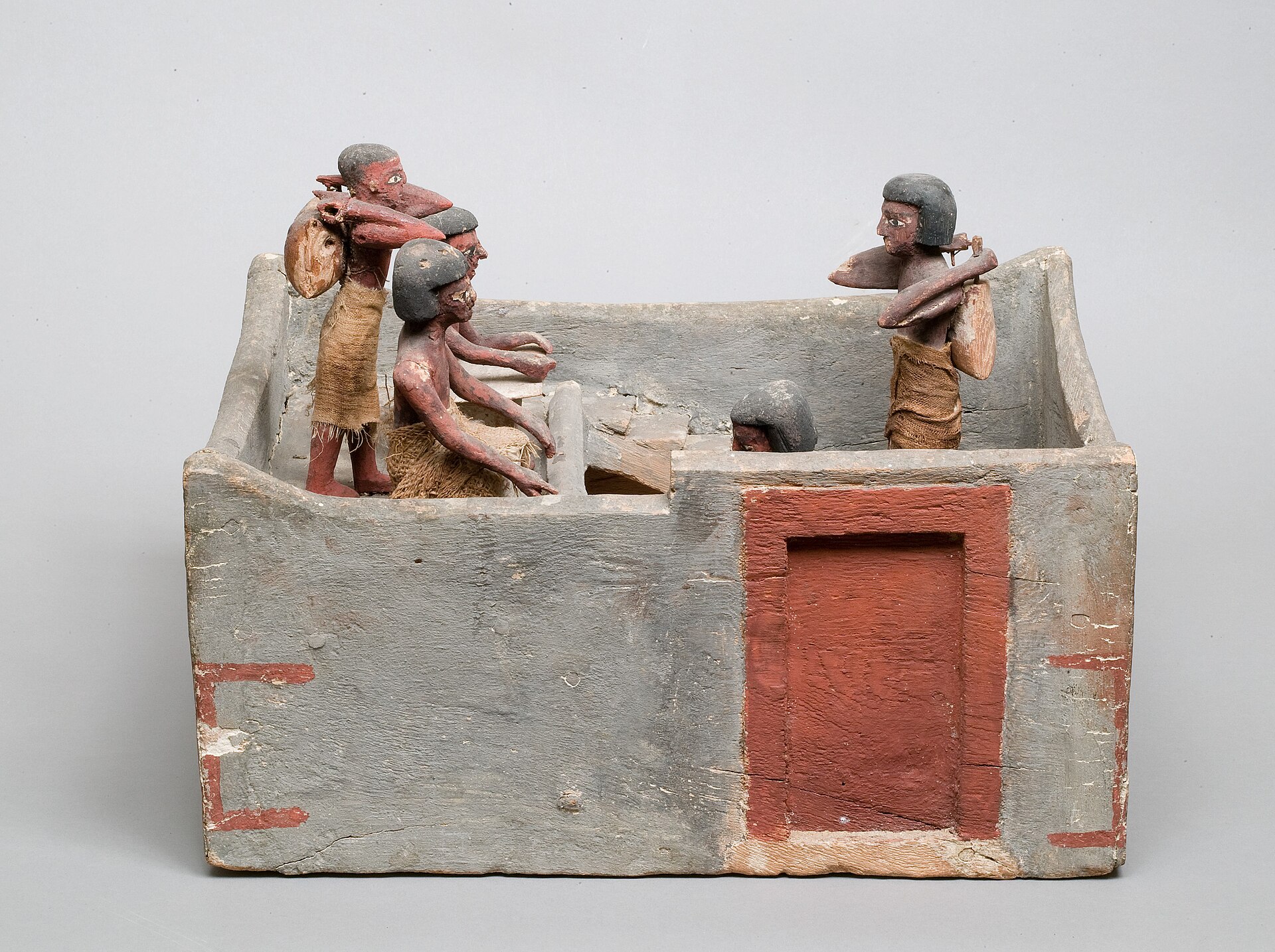 A painted wooden model of an Egyptian granary, with small figures carrying sacks of grain and a seated scribe