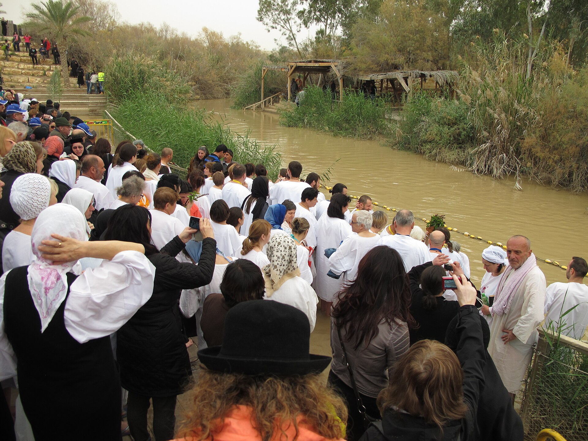 A narrow, muddy stretch of the Jordan River lined with reeds, with a large modern crowd in white robes gathered on the near bank