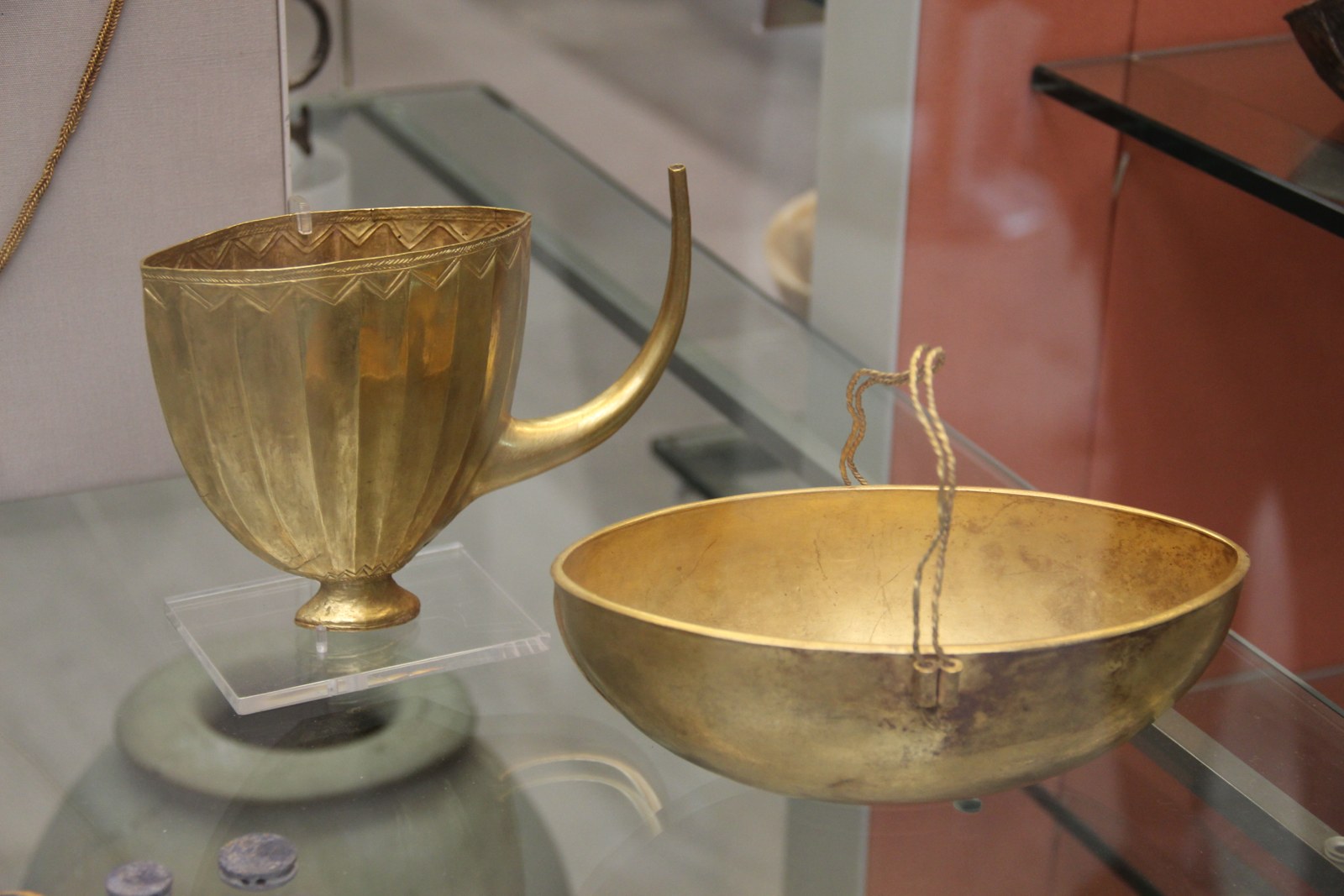 A fluted gold cup with a long curved spout beside a plain gold bowl, displayed in a museum case